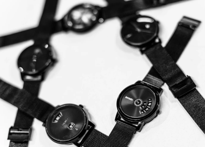 Four black wristwatches with mesh bands arranged in a circle on a white surface